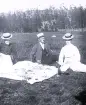 Foto av tre personer som har picknick på ute i det gröna. I bakgrunden syns skog och en gärdsgård.