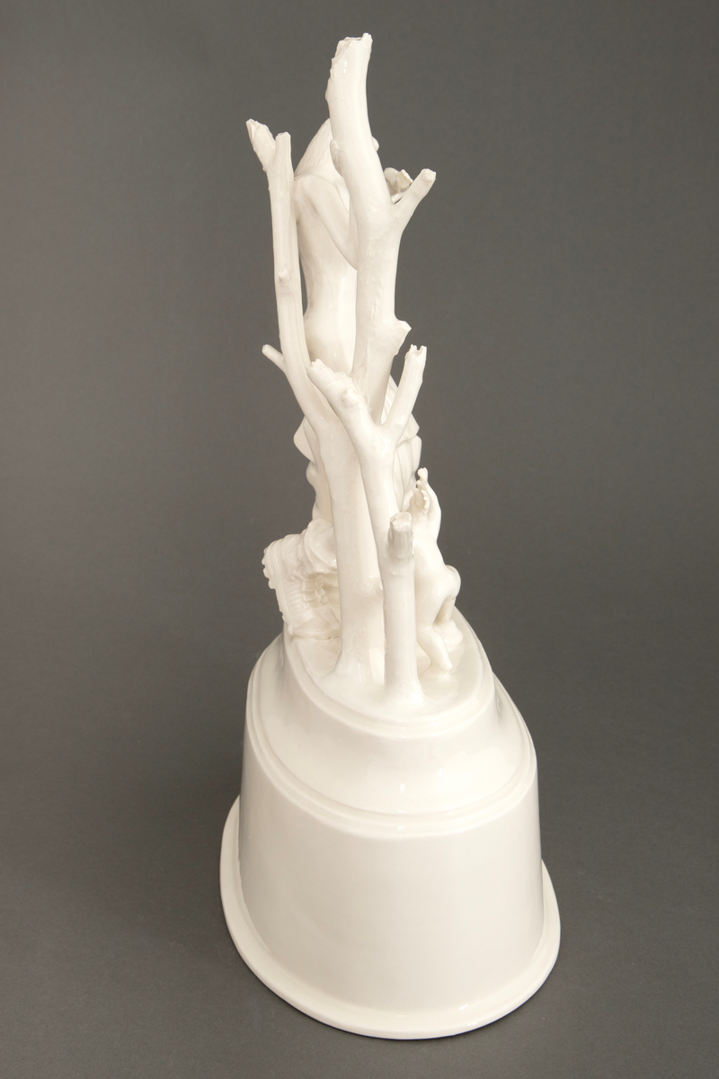 Objekt i hvitglasert porselen. På oval, hul sokkel står flere stiliserte figurer. Ytterst til høyre en figur med hestehode ikledd dress. I midten en figur med kvinnekropp og sauehode, kledd i kraftige støvler og med en avkuttet arm i ene hånden. Foran en mindre figur med hønsehode og bare en arm, vendt med ryggen mot. Ytterst til venstre to døde trær.