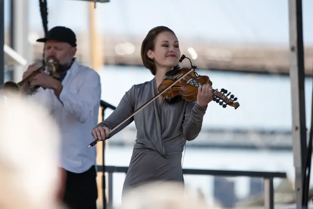 En kvinne spiller fiolin på en scene. En mann på trompet i bakgrunnen.