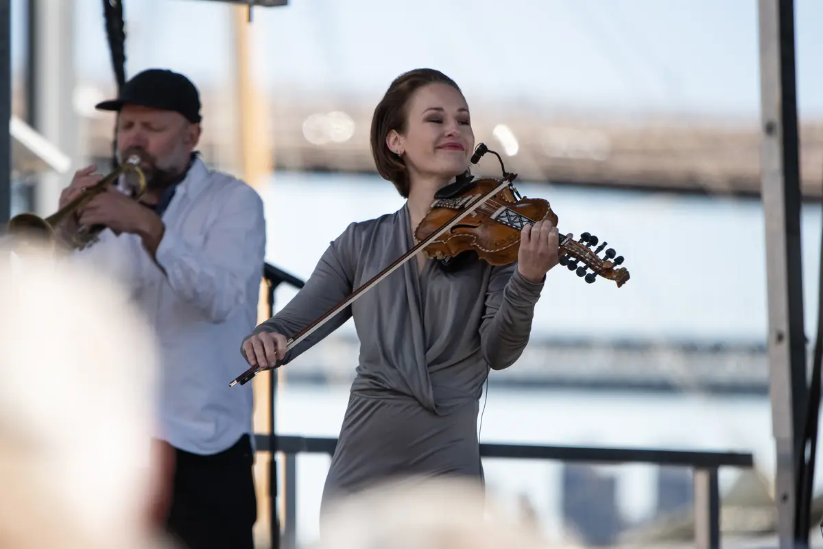 En kvinne spiller fiolin på en scene. En mann på trompet i bakgrunnen.