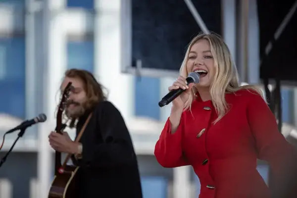 En kvinne i rødt antrekk som synger på en scene. En gitarist i bakgrunnen.