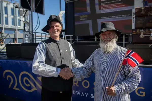 To menn med hatt og norsk flagg som tar hverandre i håndene og smiler til kameranet.