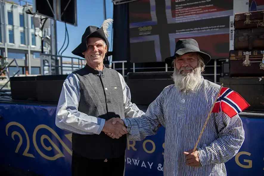 To menn med hatt og norsk flagg som tar hverandre i håndene og smiler til kameranet.