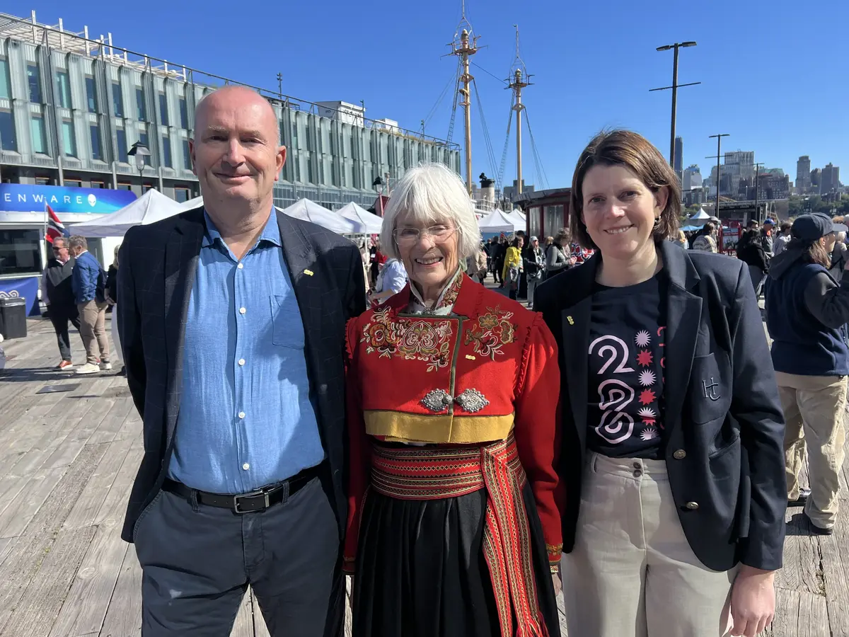 En mann og to kvinner oppstilt på en kai. De smiler til kameraet i solskinn. Kvinnen i midten har bunad, og kvinnen til høyre har Crossings 200-t-skjorte.