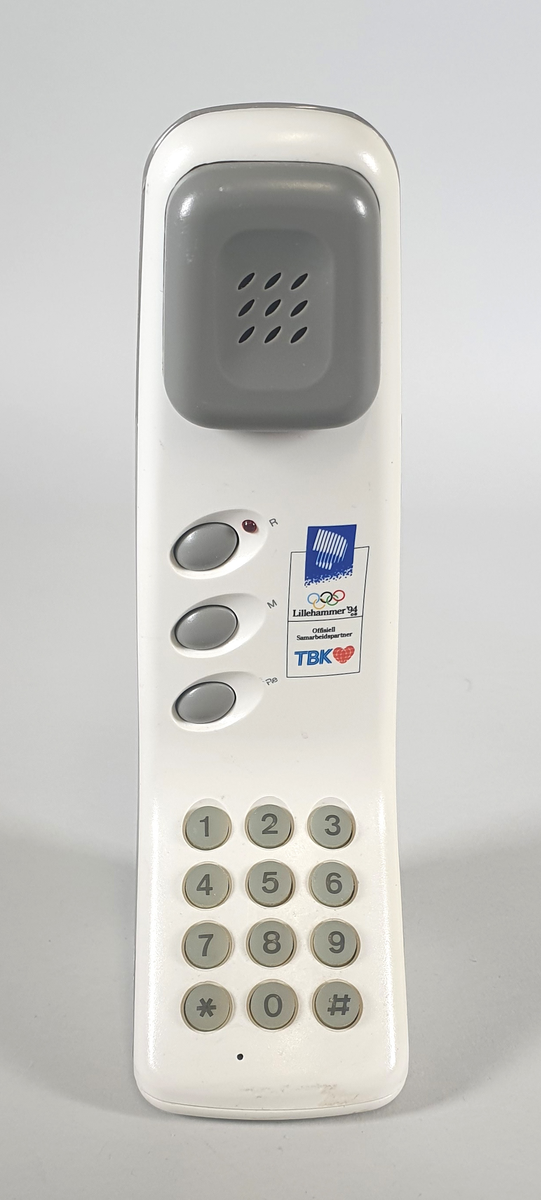 Hvit telefon med ledning, stående modell med tall-tastatur. Bred nederst, smalner inn mot toppen. Høytaler øverst. Knapp midt under for å avslutte samtalen. Logo for LIllehammer-OL og TBK over tastaturet.
