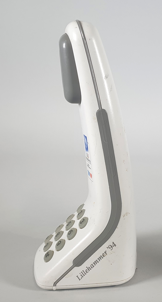 Hvit telefon med ledning, stående modell med tall-tastatur. Bred nederst, smalner inn mot toppen. Høytaler øverst. Knapp midt under for å avslutte samtalen. Logo for LIllehammer-OL og TBK over tastaturet.
