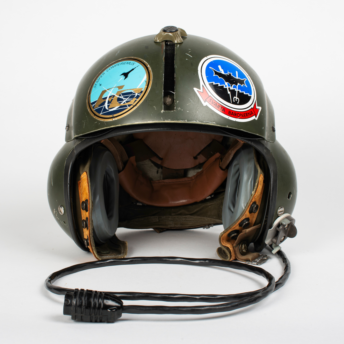 Flyghjälm SPH-4.
Grön flyghjälm med fast visirskydd av metall och tonat visir. Utksjutande form vid öronen för att ge plats för stora invändiga hörselkåpor. På vänster sida sitter en sladd för kommunikationsutrustningen, mikrofonen saknas. 
På visirskyddet sitter två runda klistermärken. Det högra med Forskningscentralens emblem med texten "SEMPER COMPERIEMUS", det vänstra är ett utbildningstecken för mörkerflygning inom marinflygning föreställande en svart fladdermus framför en blixt och en treudd samt text "SVARTA BARONERNA".