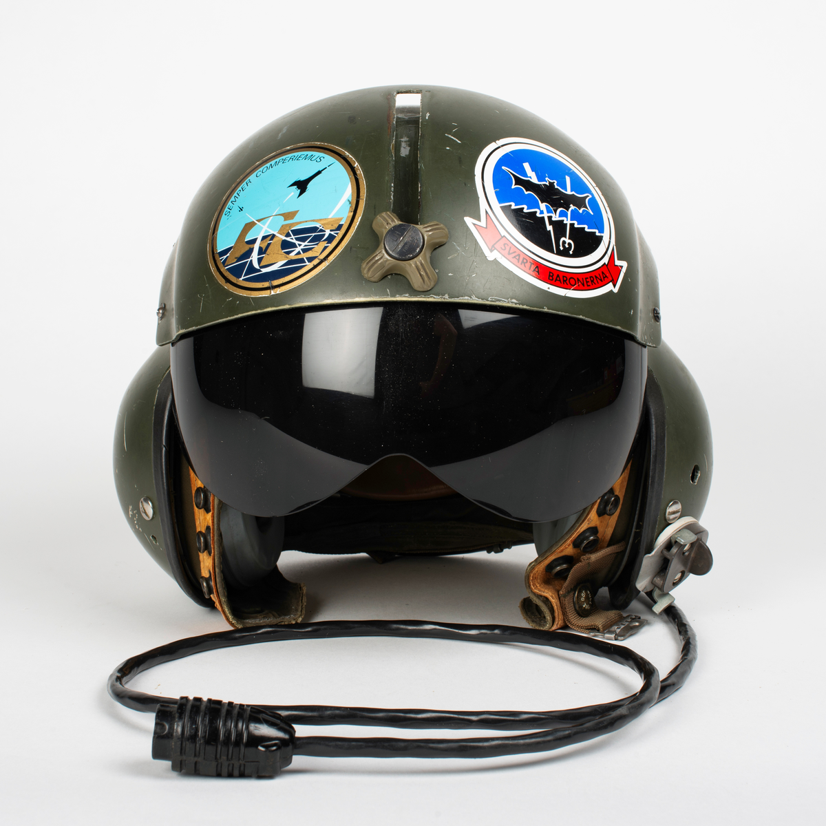 Flyghjälm SPH-4.
Grön flyghjälm med fast visirskydd av metall och tonat visir. Utksjutande form vid öronen för att ge plats för stora invändiga hörselkåpor. På vänster sida sitter en sladd för kommunikationsutrustningen, mikrofonen saknas. 
På visirskyddet sitter två runda klistermärken. Det högra med Forskningscentralens emblem med texten "SEMPER COMPERIEMUS", det vänstra är ett utbildningstecken för mörkerflygning inom marinflygning föreställande en svart fladdermus framför en blixt och en treudd samt text "SVARTA BARONERNA".
