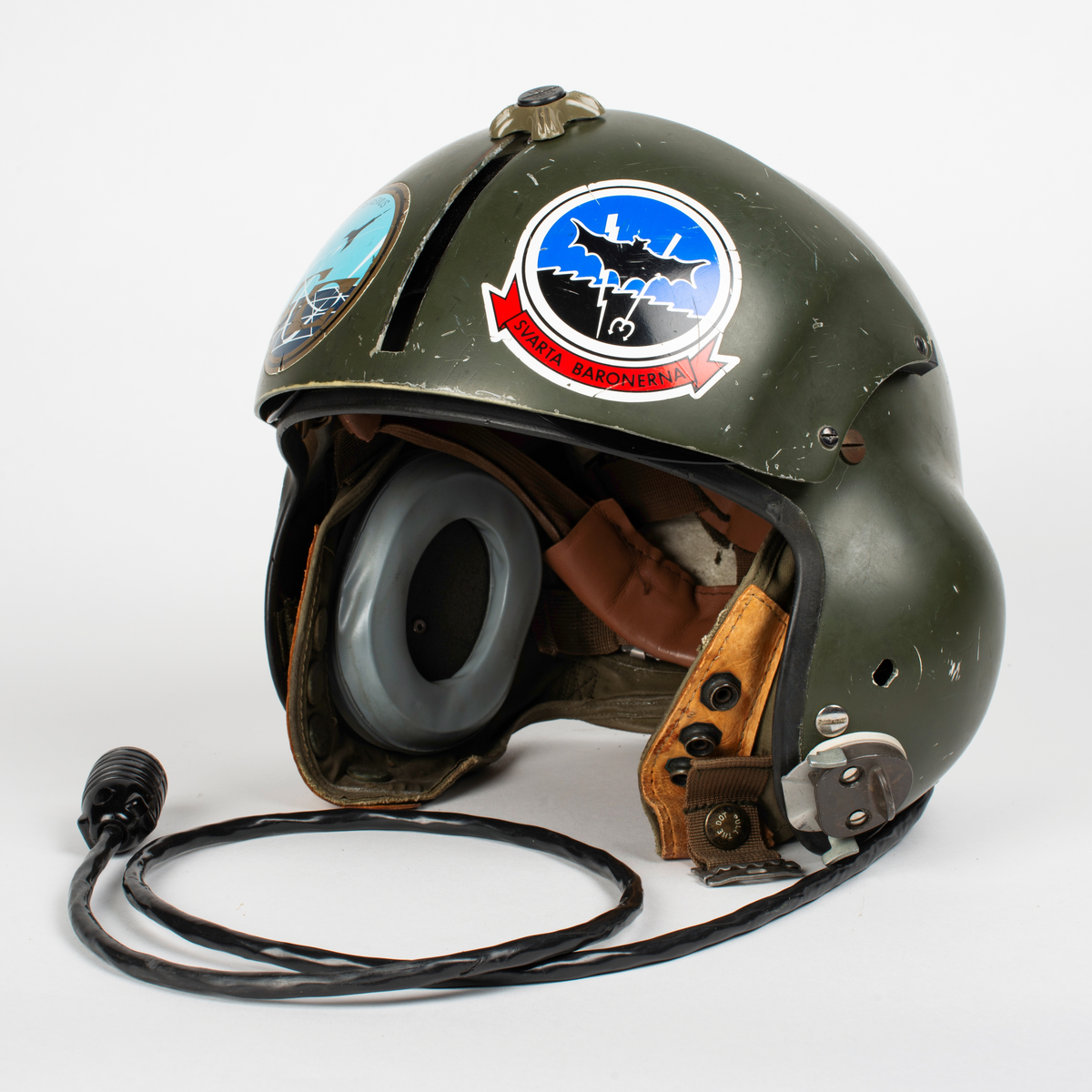 Flyghjälm SPH-4.
Grön flyghjälm med fast visirskydd av metall och tonat visir. Utksjutande form vid öronen för att ge plats för stora invändiga hörselkåpor. På vänster sida sitter en sladd för kommunikationsutrustningen, mikrofonen saknas. 
På visirskyddet sitter två runda klistermärken. Det högra med Forskningscentralens emblem med texten "SEMPER COMPERIEMUS", det vänstra är ett utbildningstecken för mörkerflygning inom marinflygning föreställande en svart fladdermus framför en blixt och en treudd samt text "SVARTA BARONERNA".