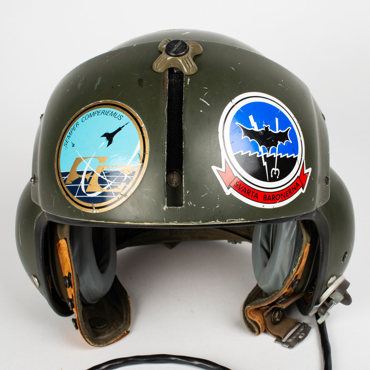 Flyghjälm SPH-4.
Grön flyghjälm med fast visirskydd av metall och tonat visir. Utksjutande form vid öronen för att ge plats för stora invändiga hörselkåpor. På vänster sida sitter en sladd för kommunikationsutrustningen, mikrofonen saknas. 
På visirskyddet sitter två runda klistermärken. Det högra med Forskningscentralens emblem med texten "SEMPER COMPERIEMUS", det vänstra är ett utbildningstecken för mörkerflygning inom marinflygning föreställande en svart fladdermus framför en blixt och en treudd samt text "SVARTA BARONERNA".