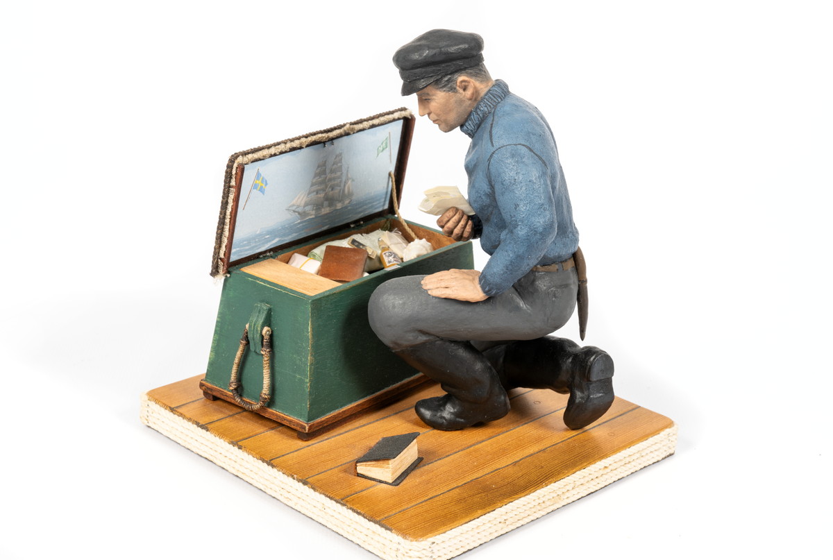 keramikfigur av en sjöman vid sin sjömanskista. Flera små detaljer i kistan. Bl a fartygsporträtt av barkskepp, målat av Hendel Friberg.