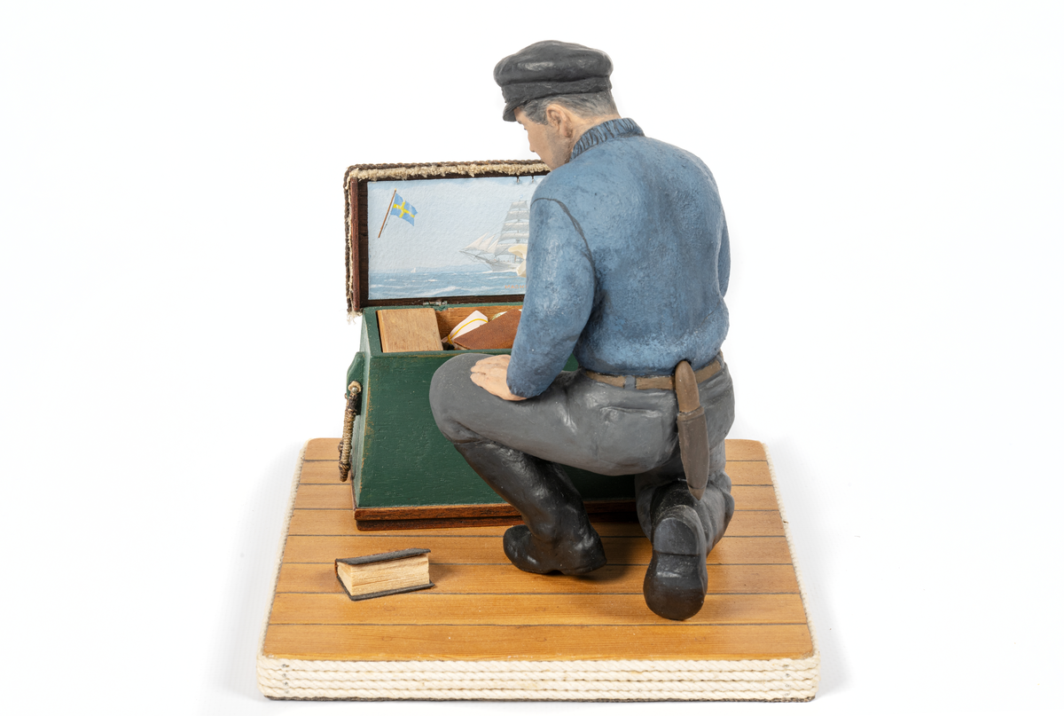 keramikfigur av en sjöman vid sin sjömanskista. Flera små detaljer i kistan. Bl a fartygsporträtt av barkskepp, målat av Hendel Friberg.