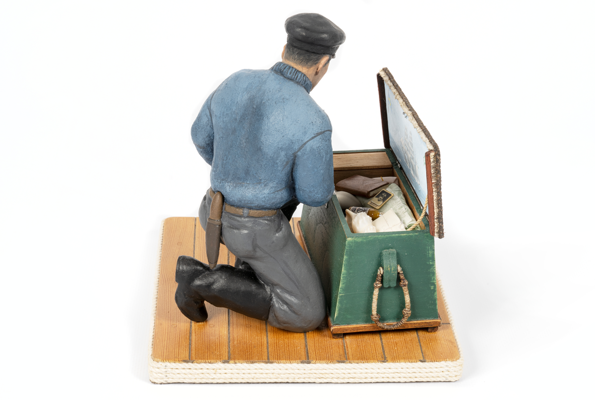 keramikfigur av en sjöman vid sin sjömanskista. Flera små detaljer i kistan. Bl a fartygsporträtt av barkskepp, målat av Hendel Friberg.