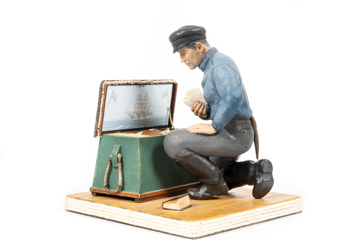 keramikfigur av en sjöman vid sin sjömanskista. Flera små detaljer i kistan. Bl a fartygsporträtt av barkskepp, målat av Hendel Friberg.