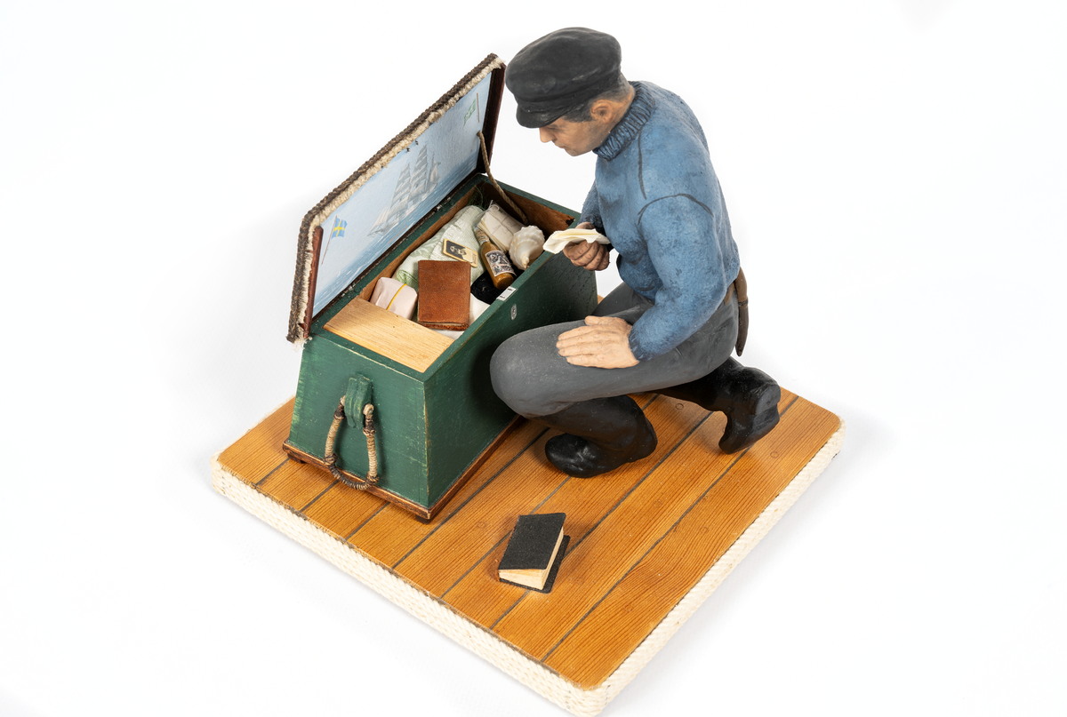 keramikfigur av en sjöman vid sin sjömanskista. Flera små detaljer i kistan. Bl a fartygsporträtt av barkskepp, målat av Hendel Friberg.