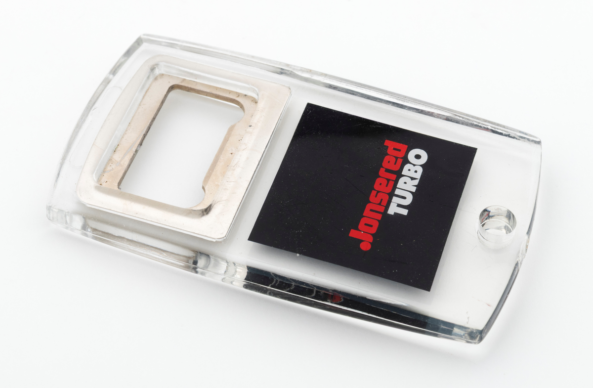 En tilnærmet rektangulær flaskeåpner som hovedsakelig er utført i støpt plast, antatt pleksiglass, med metalltann og påtrykt dekor. Metalltanna, som er støpt inni plasten/pleksiglasset har en rektangulær form. Metalltanna har en "lysåpning" som er cirka 2,6 centimeter lang og 1,4 centimeter bred. Nederst på flaskeåpeneren er det et lite hull slik at den kan henges opp eller festes til en nøkkelreing. 

Under metalltanna er det trykt et bilde av en Jonsered motorsag på den en siden og teksten Jonsered Turbo på den andre siden. Det antas at motorsagmodellen som er avbildet på flaskeåpnerens ene side skal forestille Jonsereds sagmodell 2051 Turbo, men det er ikke bekreftet. 

Flaskeåpneren er en reklameartikkel, et profileringsprodukt, for motorsagmerket Jonsered. Sagmodellen Jonsered 2051 Turbo ble lansert i 1988 og produsert til 1993. Det antas derfor at reklameartikkelen, pofileringsproduktet, ble lansert når disse sagene kom på markedet i 1988.