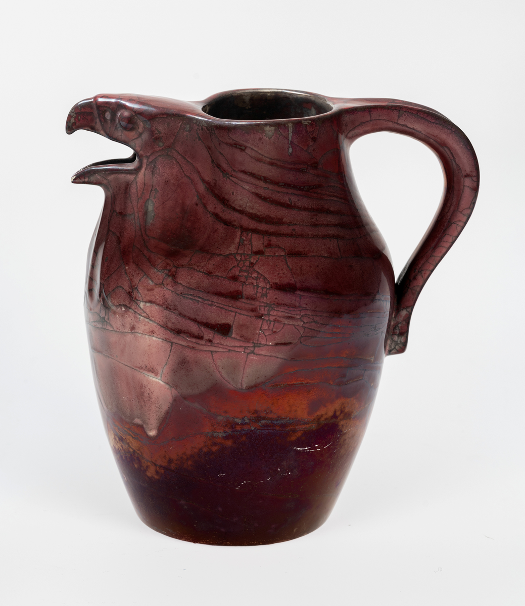 Kanne i glasert leirgods med hank. Tilnærmet ovalformet korpus med sirkulær åpning, hvor kannens tut er utformet som et skulpturelt haukehode. Overflaten er dekket med en profyrfarget glasur med store karakeleringer, hvor enkelte partier er irisert mens andre er hvite.