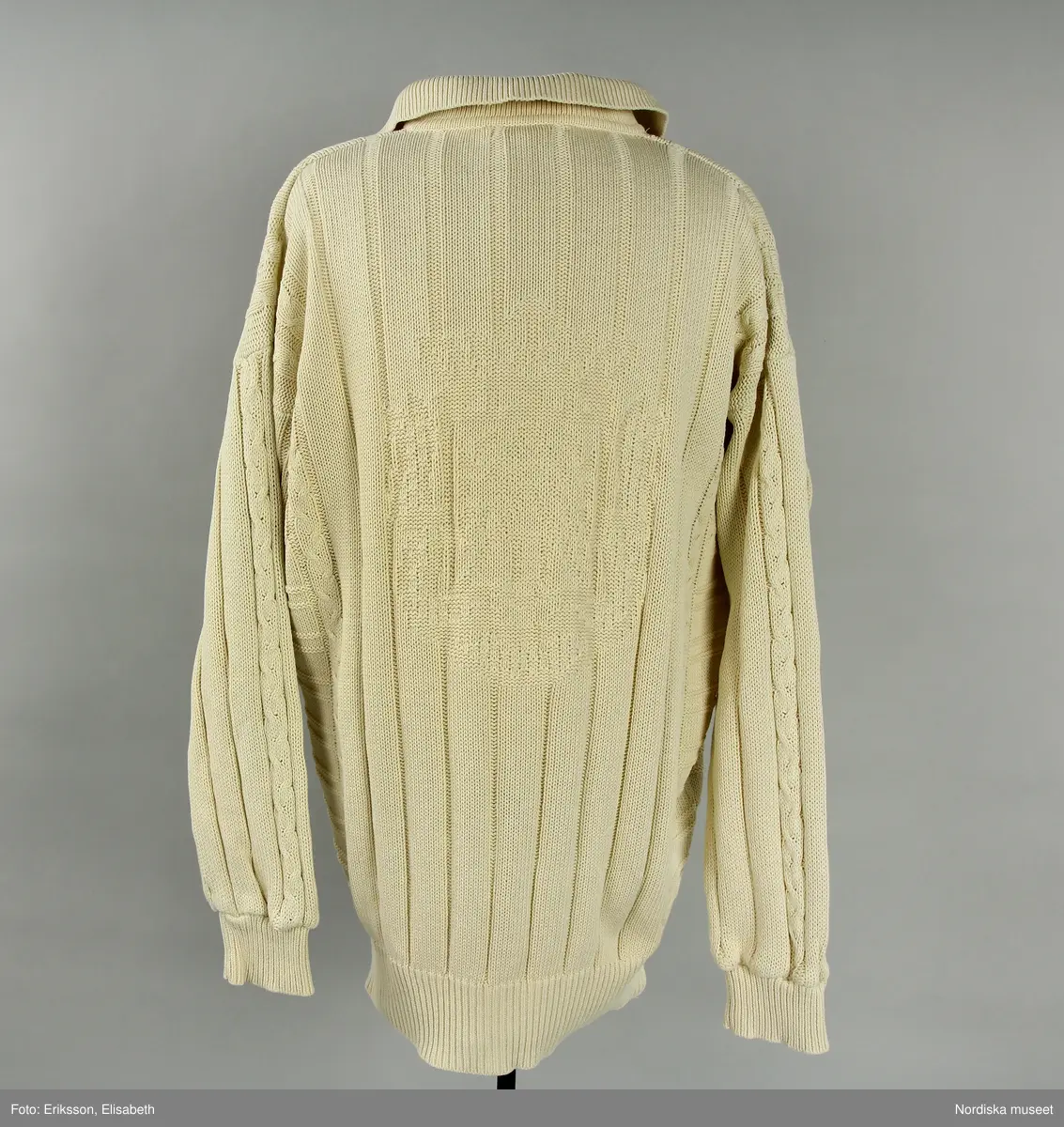 Tröja, vit, av 100% bomull. Mönsterstickad med ränder, flätor och på bakstycket vapensköld med ett B i mitten (B=Björn Borg). Ribbstickad krage samt ärmmuddar och midjemudd. Knäppning upptill fram med tre knappar av vit plast. 
I nackens insida etikett med text "BJÖRN BORG / COLLECTION / DESIGN ROHDI HEINTZ" samt logga och storleksangivelse "M". I vänster sidsöm etikett med materialdeklaration, tvättråd samt tillverkningland "Hong Kong". 
/Leif Wallin 2024-07-10