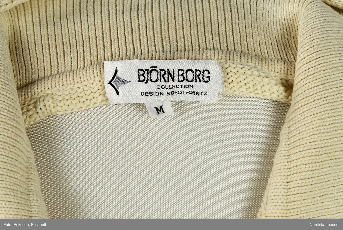 Tröja, vit, av 100% bomull. Mönsterstickad med ränder, flätor och på bakstycket vapensköld med ett B i mitten (B=Björn Borg). Ribbstickad krage samt ärmmuddar och midjemudd. Knäppning upptill fram med tre knappar av vit plast. 
I nackens insida etikett med text "BJÖRN BORG / COLLECTION / DESIGN ROHDI HEINTZ" samt logga och storleksangivelse "M". I vänster sidsöm etikett med materialdeklaration, tvättråd samt tillverkningland "Hong Kong". 
/Leif Wallin 2024-07-10