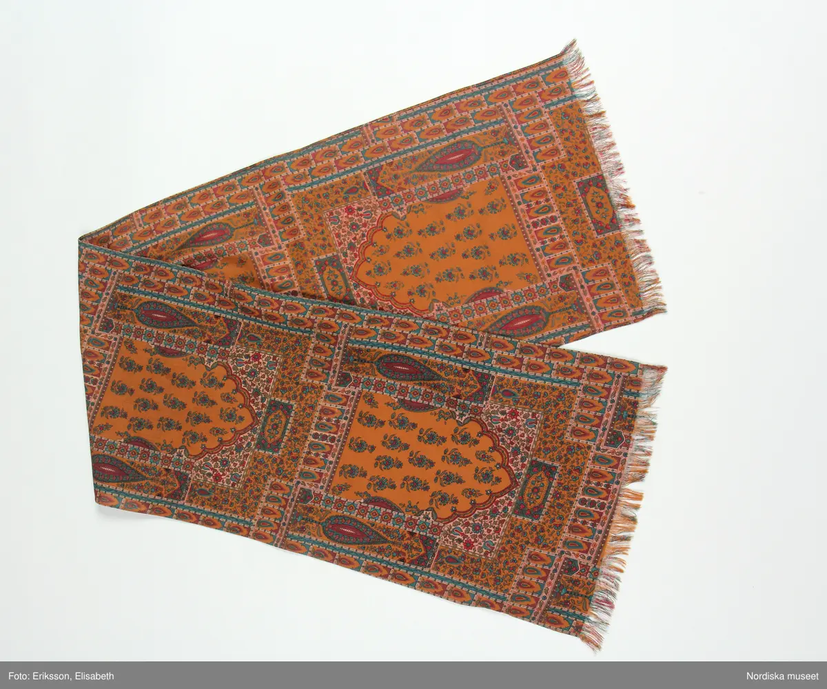 Halsduk, scarf av 100% ylle. Avlång med frans i ändarna. Tryckt orientliskt mönster (bönenisch, livets träd mm) i olika bruna, gröna och röda nyanser mot brun botten. Fållad längs långsidorna.
på ena långsidan fäst etikett med text "BJÖRN BORG / COLLECTION / DESIGN ROHDI HEINTZ" och logga samt ytterligare etikett med text "PURA LANA / MADE IN ITALY.  
/Leif Wallin 2024-07-10