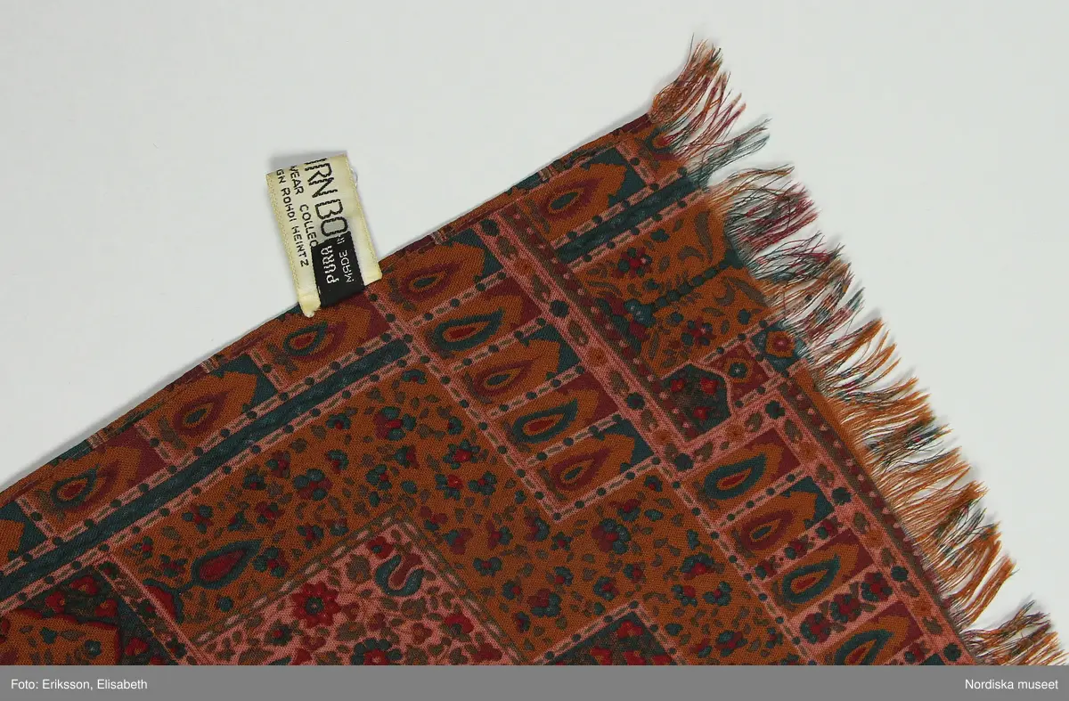 Halsduk, scarf av 100% ylle. Avlång med frans i ändarna. Tryckt orientliskt mönster (bönenisch, livets träd mm) i olika bruna, gröna och röda nyanser mot brun botten. Fållad längs långsidorna.
på ena långsidan fäst etikett med text "BJÖRN BORG / COLLECTION / DESIGN ROHDI HEINTZ" och logga samt ytterligare etikett med text "PURA LANA / MADE IN ITALY.  
/Leif Wallin 2024-07-10