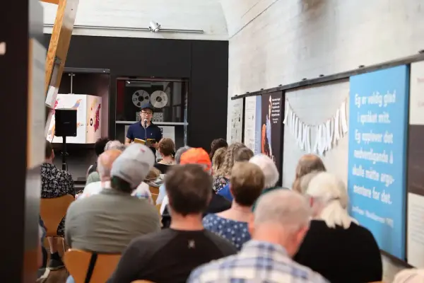 Brynjulf Jung Tjønn les høgt frå ei av bøkene sine i utstillinga i Aasentunet. I framgrunnen ser vi publikum som sit og høyrer på høgtlesinga.