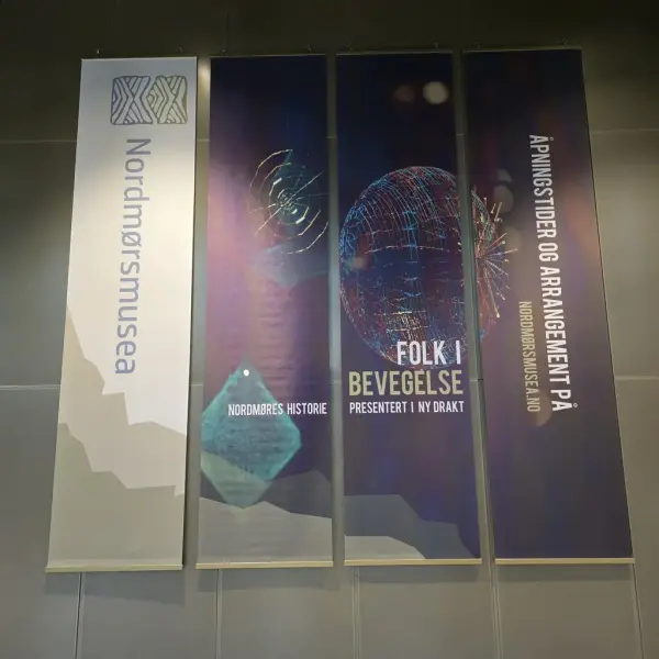 Fire vertikale bannere på en vegg. Første banner viser 'Nordmørsmusea' med logo. Andre banner har teksten 'NORDMØRES HISTORIE' med abstrakt design. Tredje banner viser en wireframe-sfære og teksten 'FOLK I BEVEGELSE PRESENTERT I NY DRAKT'. Fjerde banner informerer om åpningstider og arrangementer på nordmorsmusea.no.