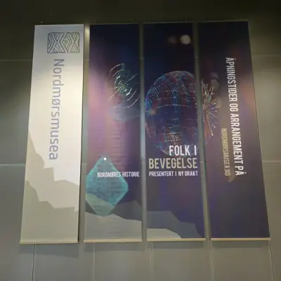 Fire vertikale bannere på en vegg. Første banner viser 'Nordmørsmusea' med logo. Andre banner har teksten 'NORDMØRES HISTORIE' med abstrakt design. Tredje banner viser en wireframe-sfære og teksten 'FOLK I BEVEGELSE PRESENTERT I NY DRAKT'. Fjerde banner informerer om åpningstider og arrangementer på nordmorsmusea.no.