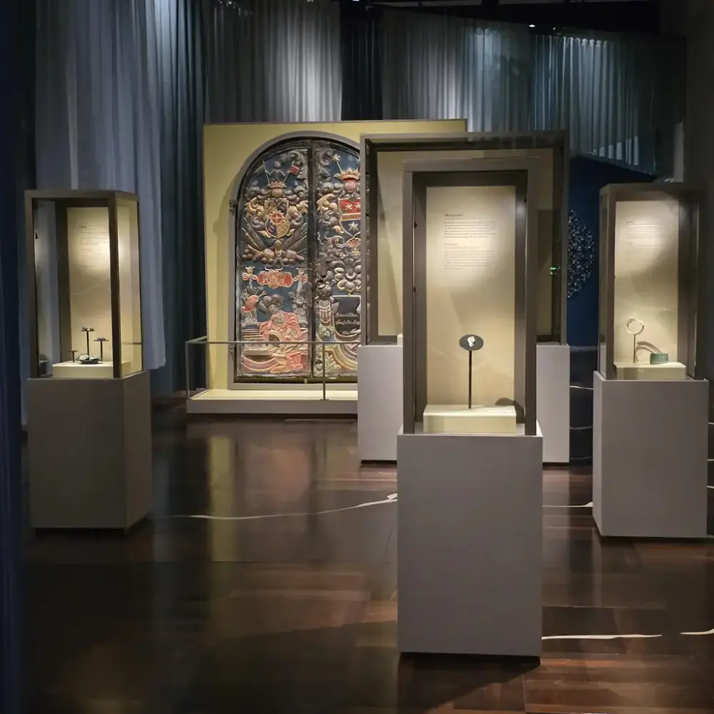 Et utstillingsrom med flere monterte glasskasser som inneholder belyste gjenstander. I bakgrunnen sees et stort dekorativt panel med utskjæringer og fargerike detaljer.