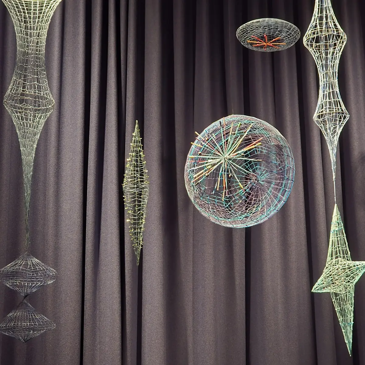  Fargerike og intrikate wireframe-skulpturer henger foran et mørkt sceneteppe. Skulpturene har geometriske og abstrakte former, inkludert sfærer og symmetriske design, og fremstår som belyste eller reflekterende.