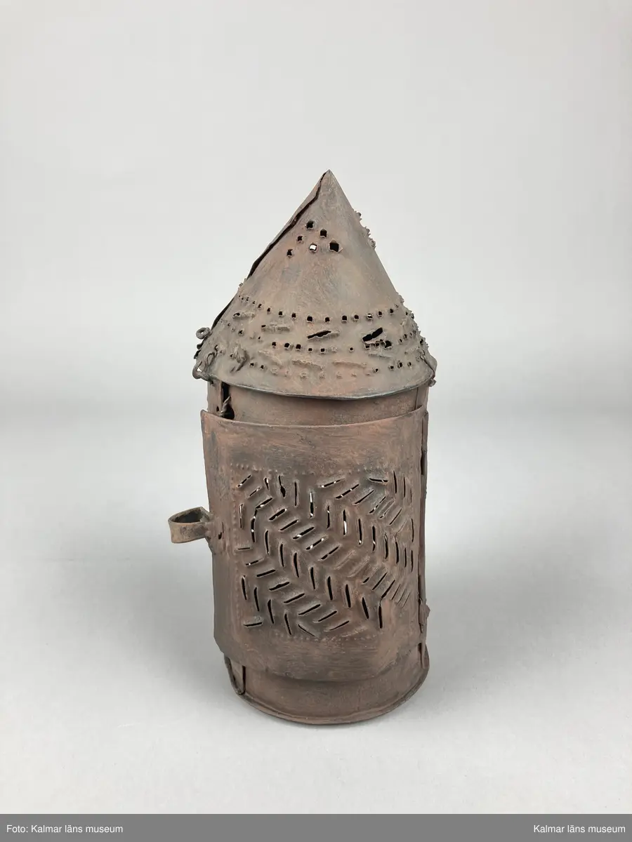 KLM 46903. Lykta, handlykta. Av metall med genombrutet mönster. Rund form med konisk överdel. Dörr på sidan. En ljushållare. Målad i brun rostskyddsfärg.