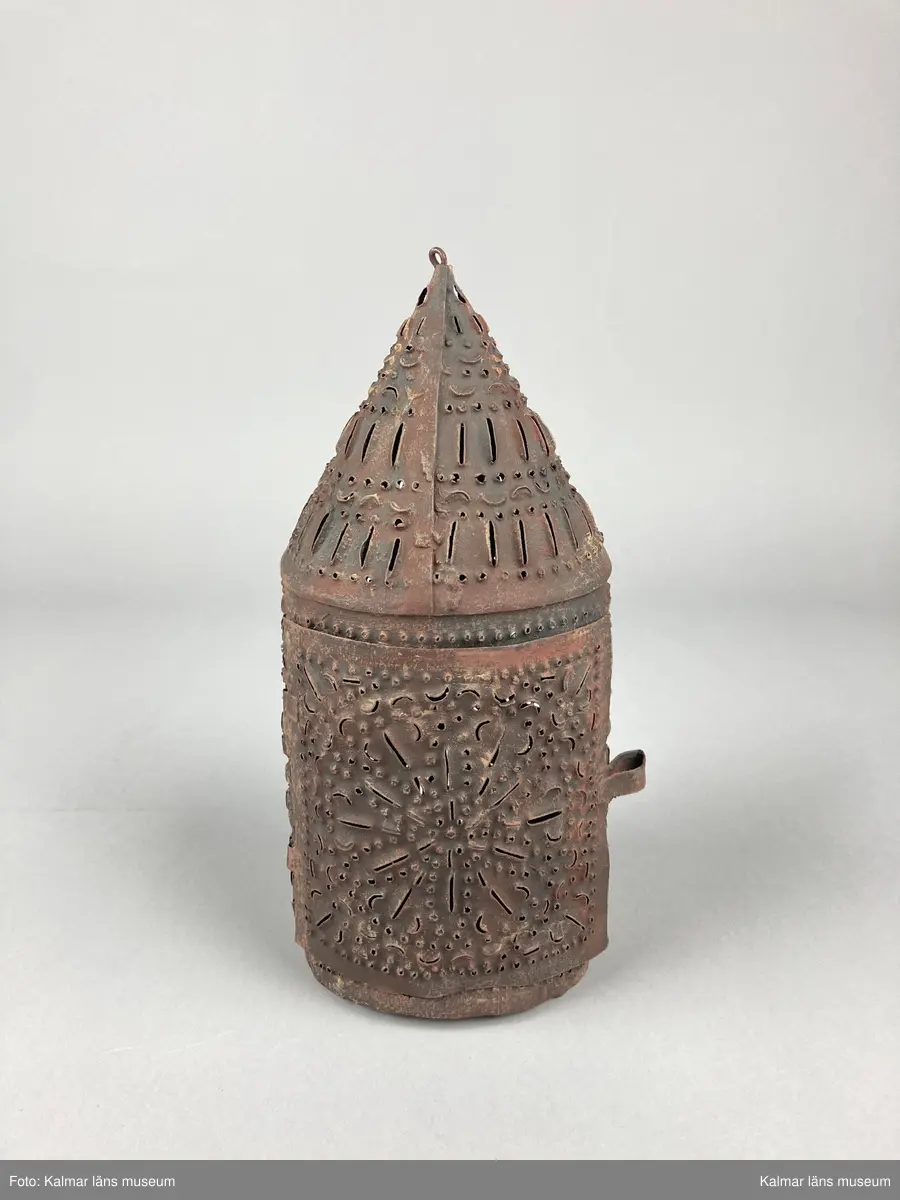 KLM 46904. Lykta, handlykta. Av metall med genombrutet mönster. Rund form med konisk överdel. Dörr på sidan. En ljushållare. Målad i brun rostskyddsfärg.