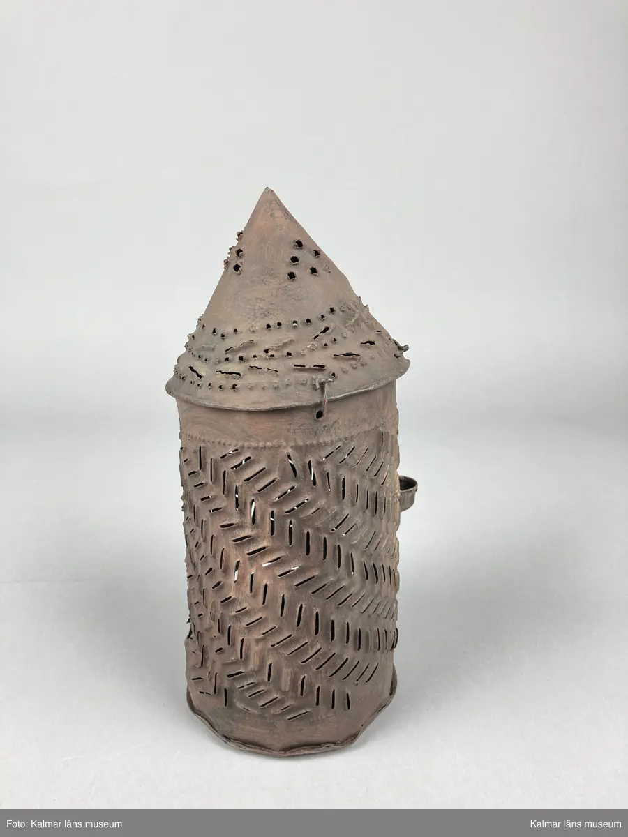 KLM 46903. Lykta, handlykta. Av metall med genombrutet mönster. Rund form med konisk överdel. Dörr på sidan. En ljushållare. Målad i brun rostskyddsfärg.