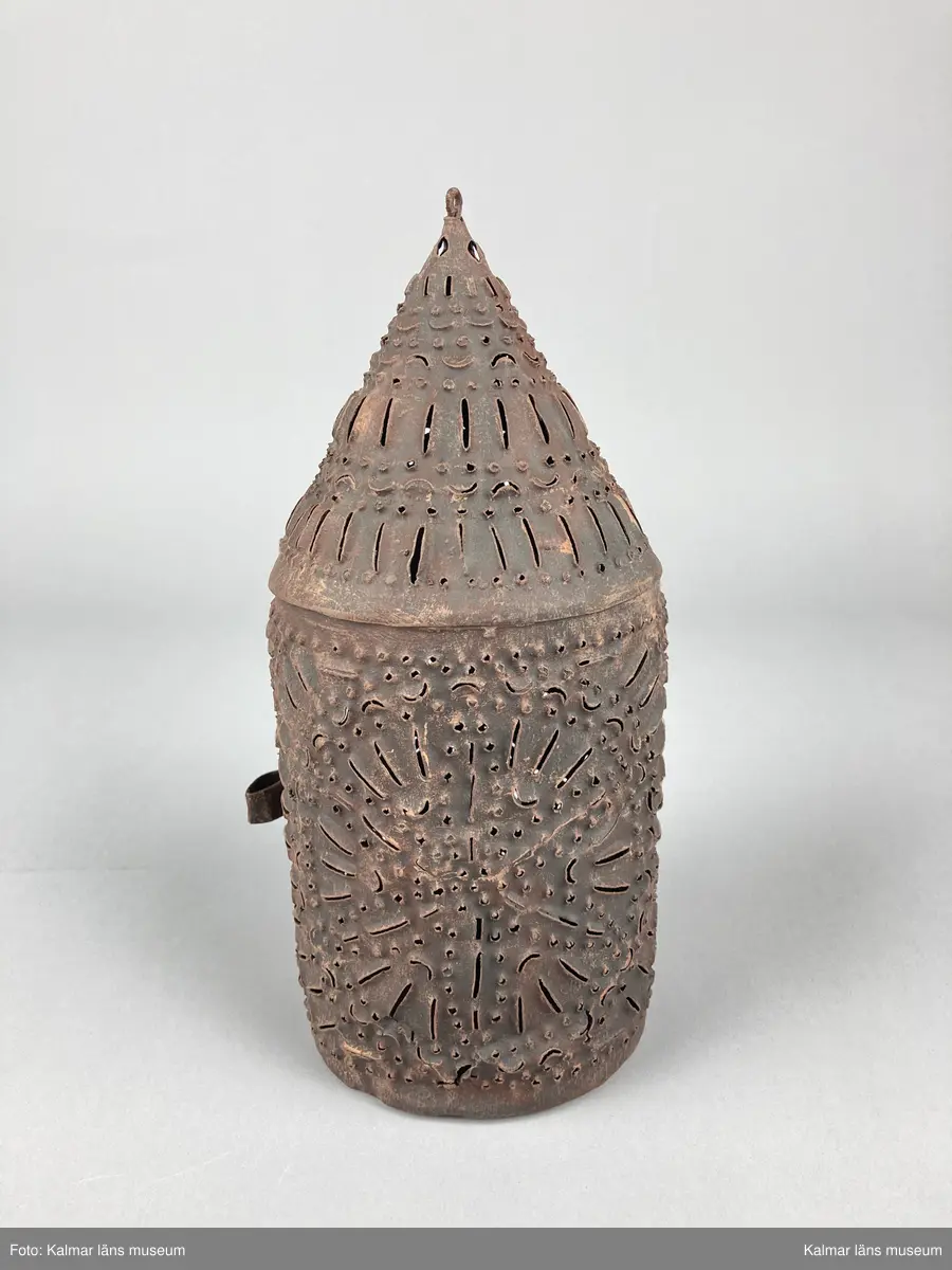 KLM 46904. Lykta, handlykta. Av metall med genombrutet mönster. Rund form med konisk överdel. Dörr på sidan. En ljushållare. Målad i brun rostskyddsfärg.