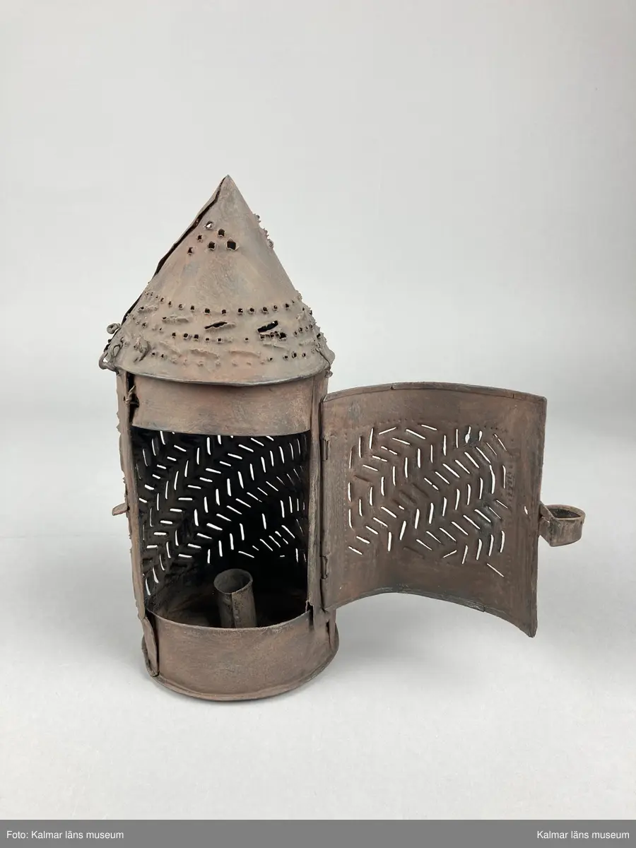 KLM 46903. Lykta, handlykta. Av metall med genombrutet mönster. Rund form med konisk överdel. Dörr på sidan. En ljushållare. Målad i brun rostskyddsfärg.