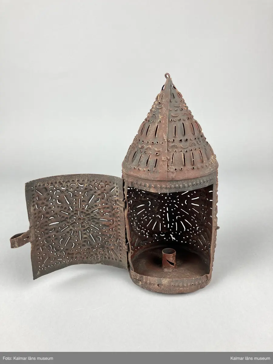 KLM 46904. Lykta, handlykta. Av metall med genombrutet mönster. Rund form med konisk överdel. Dörr på sidan. En ljushållare. Målad i brun rostskyddsfärg.