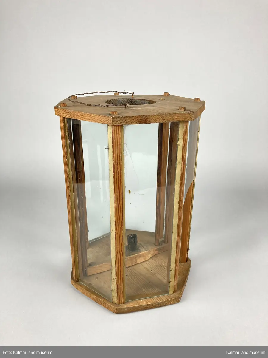 KLM 46961. Lykta, handlykta. Av trä. Sjusidig med glasrutor. Plan ovansida med centrerat hål och handtag av tvinnad metalltråd. Invändigt med en ljushållare. Tändning av ljus sker genom att ljushållaren lyftes upp med en på ena sidan fastsatt stav genom ovansidans hål. Tre av glasrutorna är trasiga.