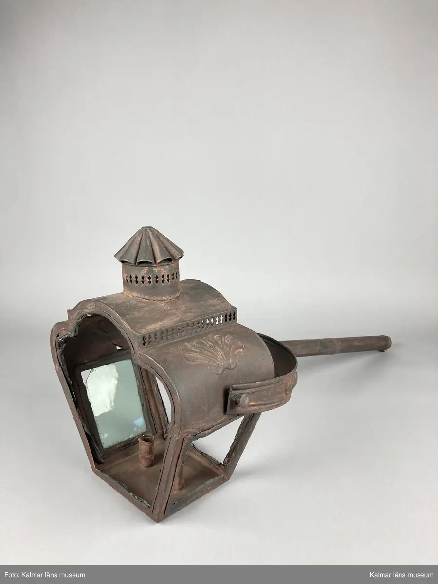 KLM 46962. Lykta, stånglykta. Av järnbleck och glas. Fyrsidigt lykthus med rektangulär bottenplatta och två ljushållare, en kortsida öppningsbar. Tre sidor utan glas. Via kortsidorna sammansatt med ett rörligt handtag med gaffelform. Handtaget med delvis räfflat mönster och förvaring. Lykthusets överdel profilerat med formpressat mönster i sidorna och en cirkulär kåpa. Därtill med genombrutet gallerverk. Målad i brun rostskyddsfärg.