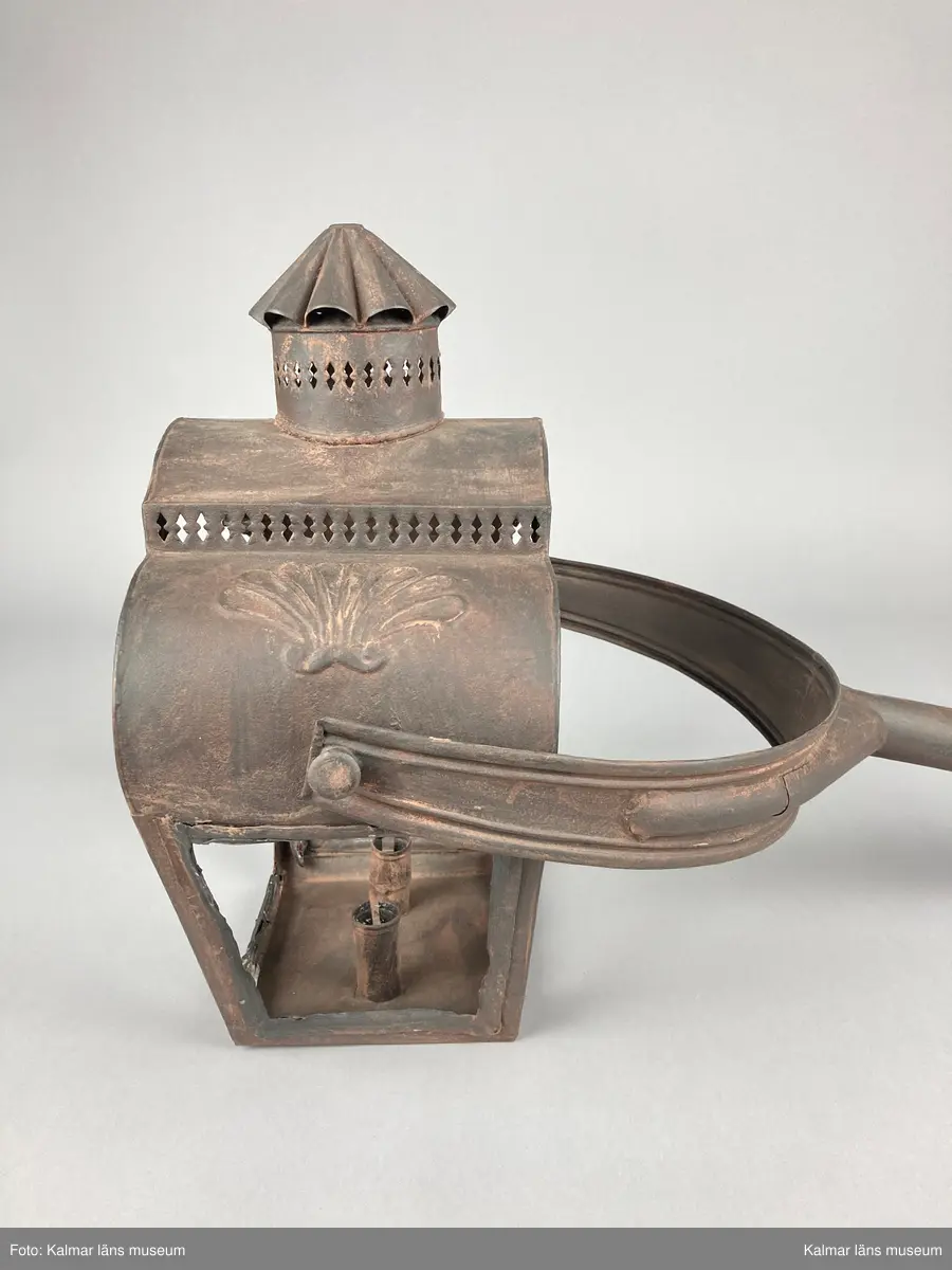 KLM 46962. Lykta, stånglykta. Av järnbleck och glas. Fyrsidigt lykthus med rektangulär bottenplatta och två ljushållare, en kortsida öppningsbar. Tre sidor utan glas. Via kortsidorna sammansatt med ett rörligt handtag med gaffelform. Handtaget med delvis räfflat mönster och förvaring. Lykthusets överdel profilerat med formpressat mönster i sidorna och en cirkulär kåpa. Därtill med genombrutet gallerverk. Målad i brun rostskyddsfärg.