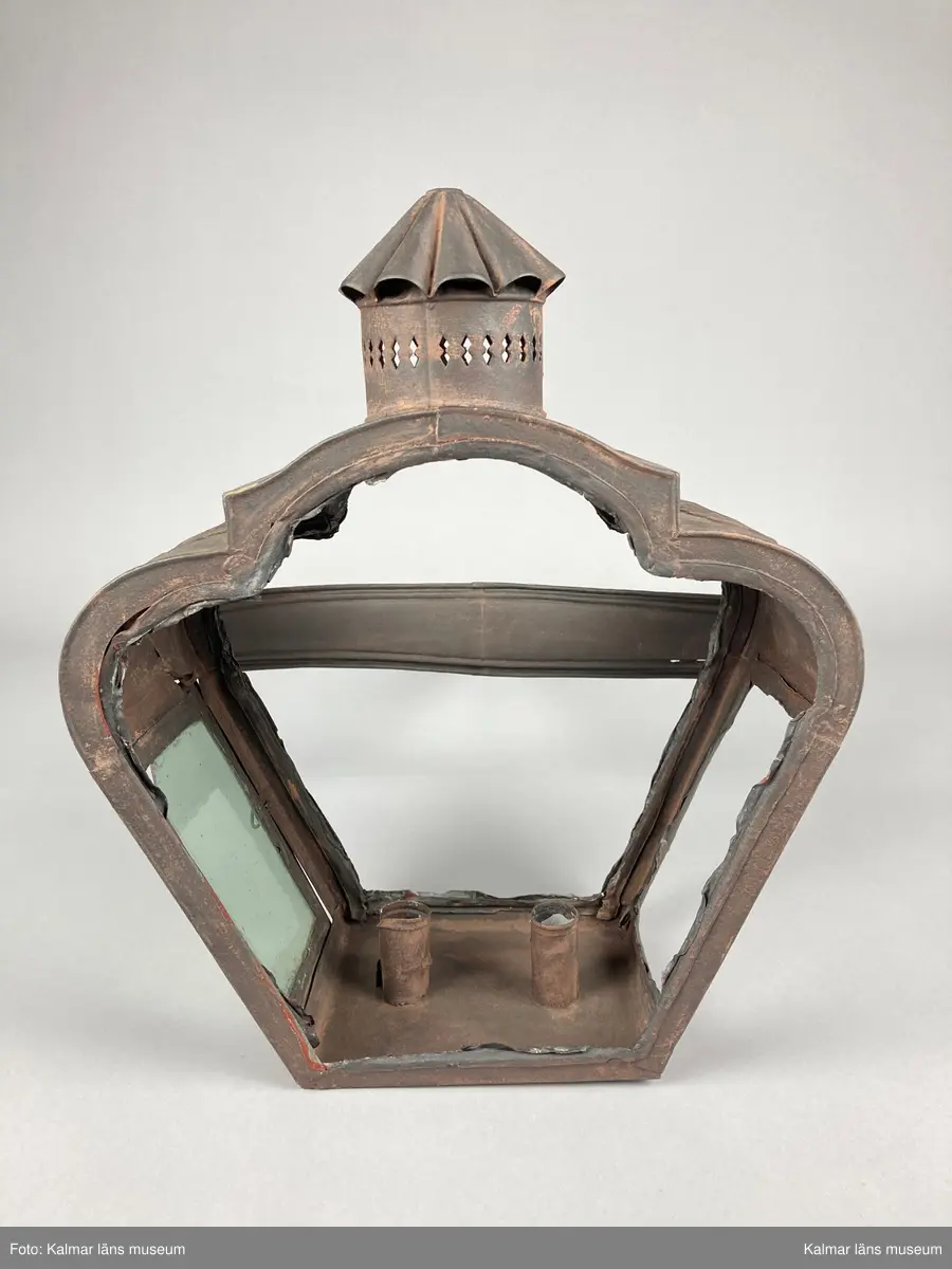 KLM 46962. Lykta, stånglykta. Av järnbleck och glas. Fyrsidigt lykthus med rektangulär bottenplatta och två ljushållare, en kortsida öppningsbar. Tre sidor utan glas. Via kortsidorna sammansatt med ett rörligt handtag med gaffelform. Handtaget med delvis räfflat mönster och förvaring. Lykthusets överdel profilerat med formpressat mönster i sidorna och en cirkulär kåpa. Därtill med genombrutet gallerverk. Målad i brun rostskyddsfärg.