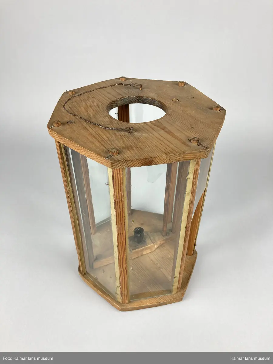 KLM 46961. Lykta, handlykta. Av trä. Sjusidig med glasrutor. Plan ovansida med centrerat hål och handtag av tvinnad metalltråd. Invändigt med en ljushållare. Tändning av ljus sker genom att ljushållaren lyftes upp med en på ena sidan fastsatt stav genom ovansidans hål. Tre av glasrutorna är trasiga.