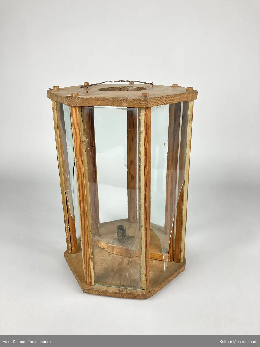 KLM 46961. Lykta, handlykta. Av trä. Sjusidig med glasrutor. Plan ovansida med centrerat hål och handtag av tvinnad metalltråd. Invändigt med en ljushållare. Tändning av ljus sker genom att ljushållaren lyftes upp med en på ena sidan fastsatt stav genom ovansidans hål. Tre av glasrutorna är trasiga.