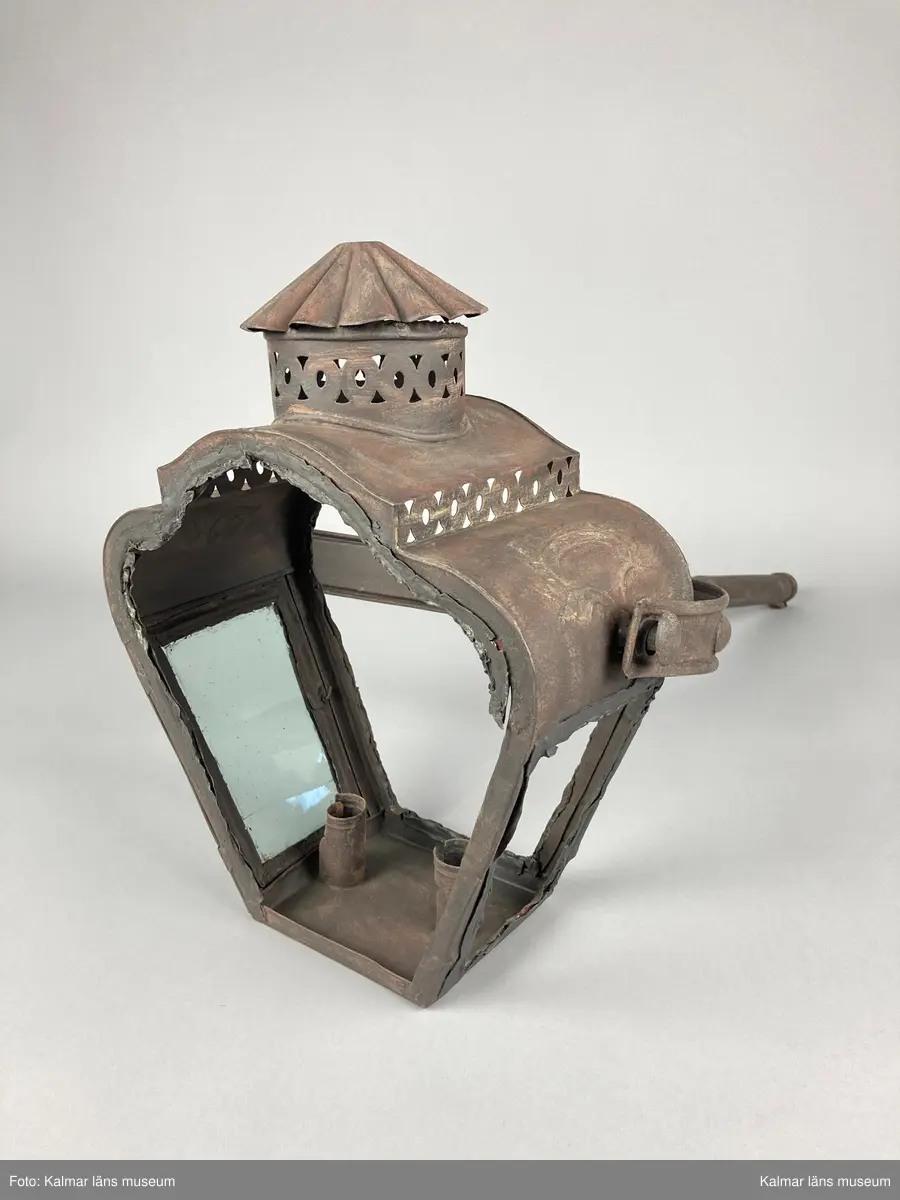 KLM 46963. Lykta, stånglykta. Av järnbleck och glas. Fyrsidigt lykthus med rektangulär bottenplatta och två ljushållare, en kortsida öppningsbar. Tre sidor utan glas. Via kortsidorna sammansatt med ett rörligt handtag med gaffelform. Handtaget med delvis räfflat mönster och förvaring. Lykthusets överdel profilerat med formpressat mönster i sidorna och en cirkulär kåpa. Därtill med genombrutet gallerverk. Målad i brun rostskyddsfärg.