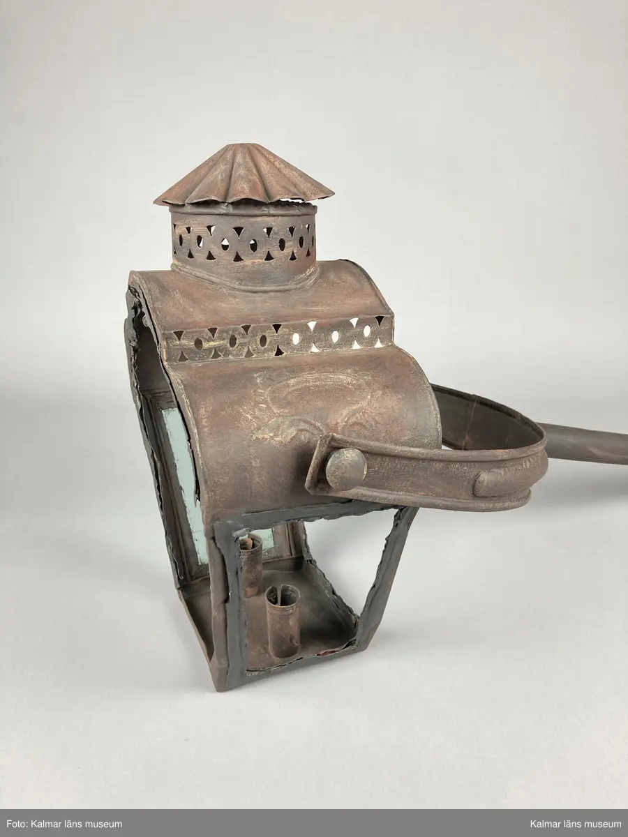 KLM 46963. Lykta, stånglykta. Av järnbleck och glas. Fyrsidigt lykthus med rektangulär bottenplatta och två ljushållare, en kortsida öppningsbar. Tre sidor utan glas. Via kortsidorna sammansatt med ett rörligt handtag med gaffelform. Handtaget med delvis räfflat mönster och förvaring. Lykthusets överdel profilerat med formpressat mönster i sidorna och en cirkulär kåpa. Därtill med genombrutet gallerverk. Målad i brun rostskyddsfärg.