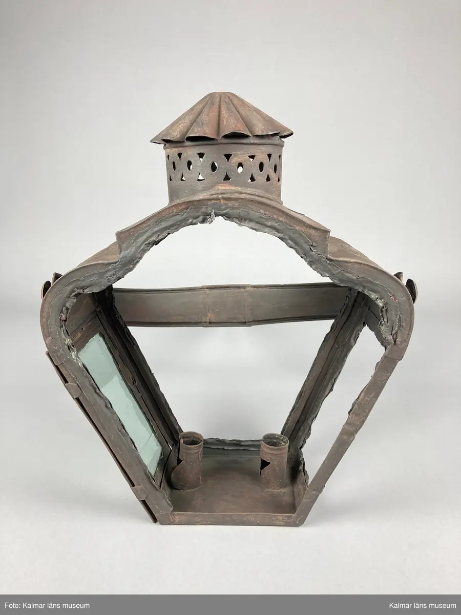 KLM 46963. Lykta, stånglykta. Av järnbleck och glas. Fyrsidigt lykthus med rektangulär bottenplatta och två ljushållare, en kortsida öppningsbar. Tre sidor utan glas. Via kortsidorna sammansatt med ett rörligt handtag med gaffelform. Handtaget med delvis räfflat mönster och förvaring. Lykthusets överdel profilerat med formpressat mönster i sidorna och en cirkulär kåpa. Därtill med genombrutet gallerverk. Målad i brun rostskyddsfärg.