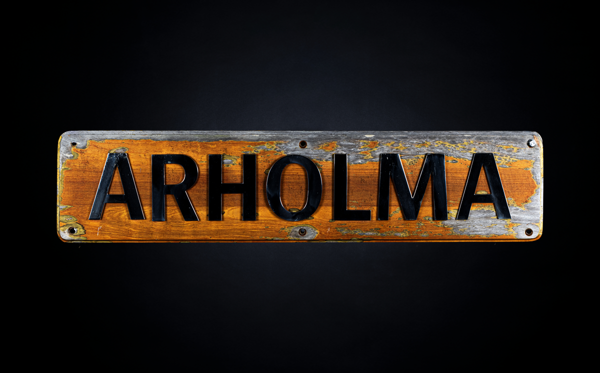 Namnbrädan är en rektangulärt formad skylt med något avrundade kanter bestående av en träplatta. På skyltens framsida ses texten "ARHOLMA" med silvriga bokstäver. På baksidan ses 22 skruvar och muttrar som fäster bokstäverna samt tre upphöjda partier för att kunna hänga upp skylten i sex skruvar.