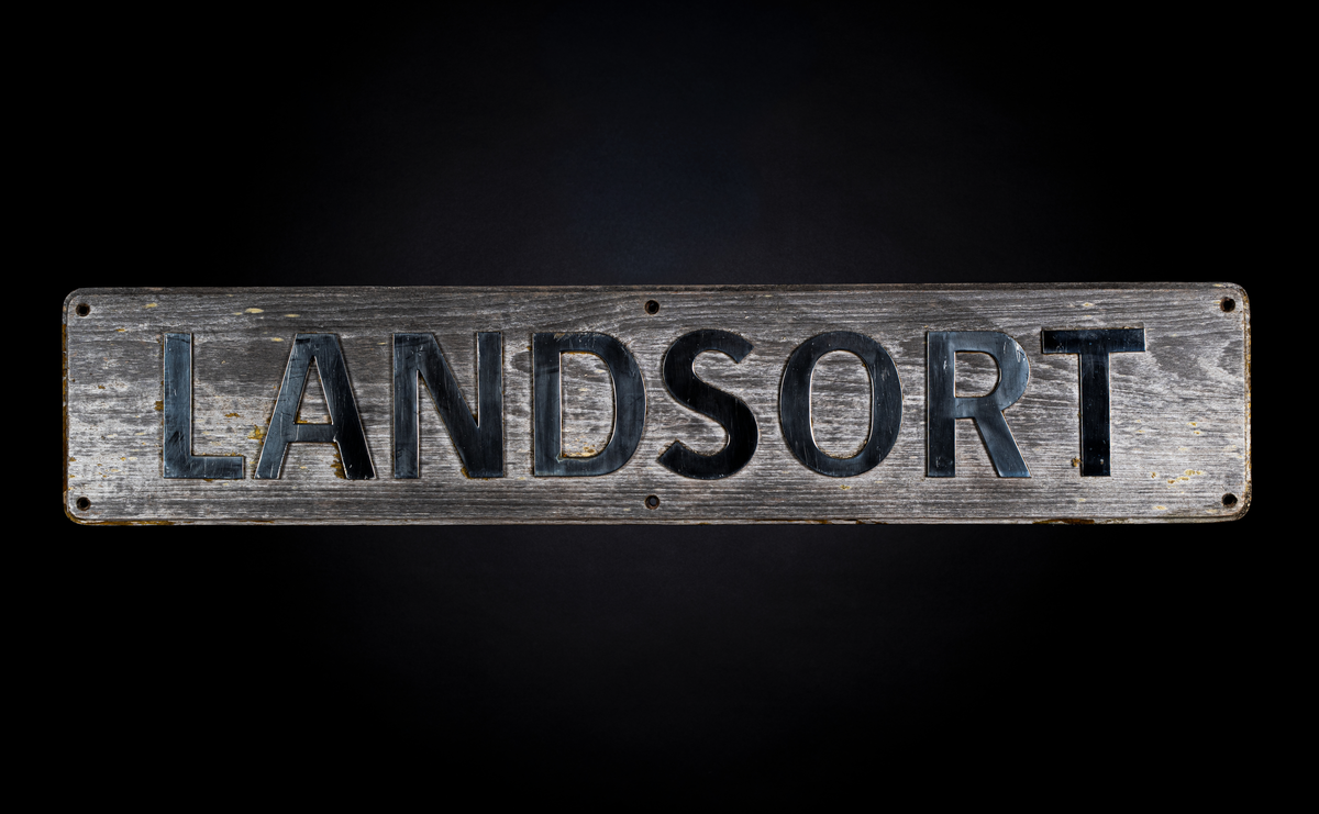 Namnbrädan är av en rektangulärt formad skylt med något avrundade kanter bestående av en träplatta. På skyltens framsida ses texten "LANDSORT" med silvriga bokstäver och sex håligheter för att kunna hänga upp den i kraftiga skruvar. På baksidan finns 23 skruvar och muttrar som fäster bokstäverna.