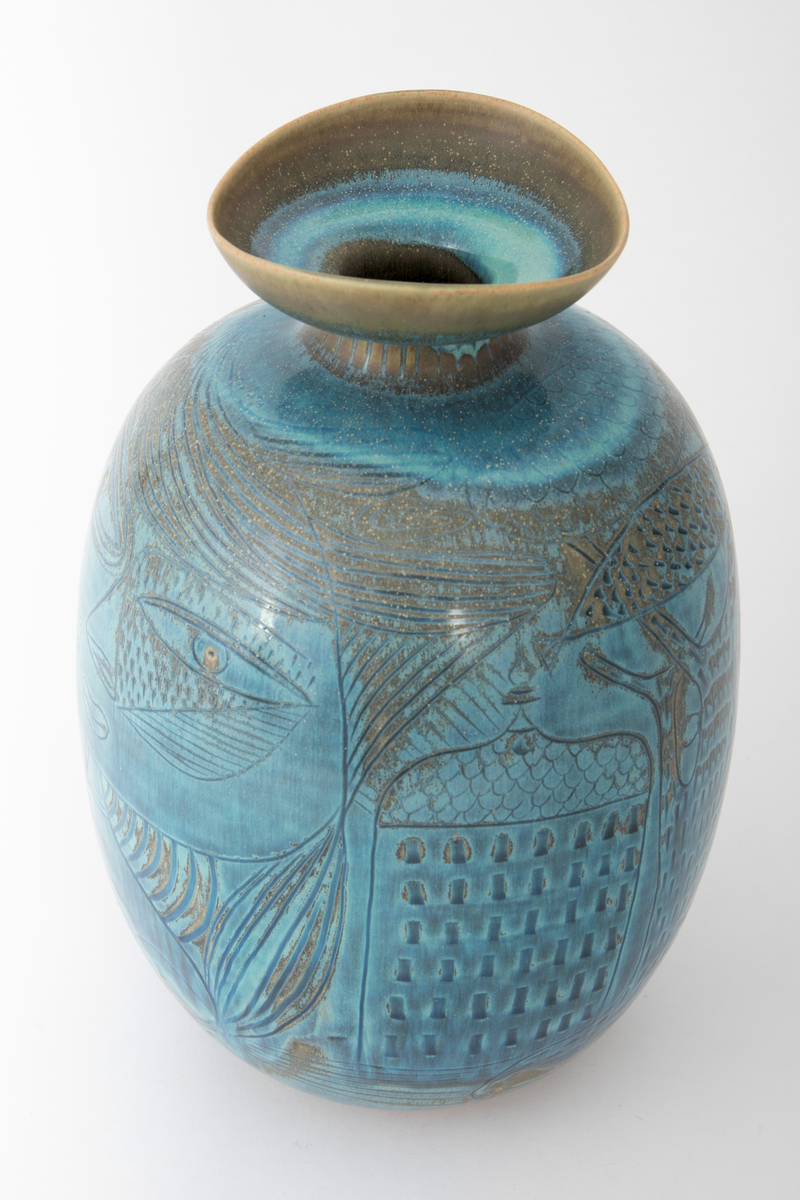 Krukke i steingods. Oval korpusform og kort innsnevret hals med vid, skålformet krage om munningen. Dekket med glasur i blå nyanser med overganger mot grågrønt. Innrisset dekor forestillende en kvinne i halvfigur, hus, tre, fisk samt en kvinne i helfigur mot en mønstret bakgrunn med ovaler og små ansikter.