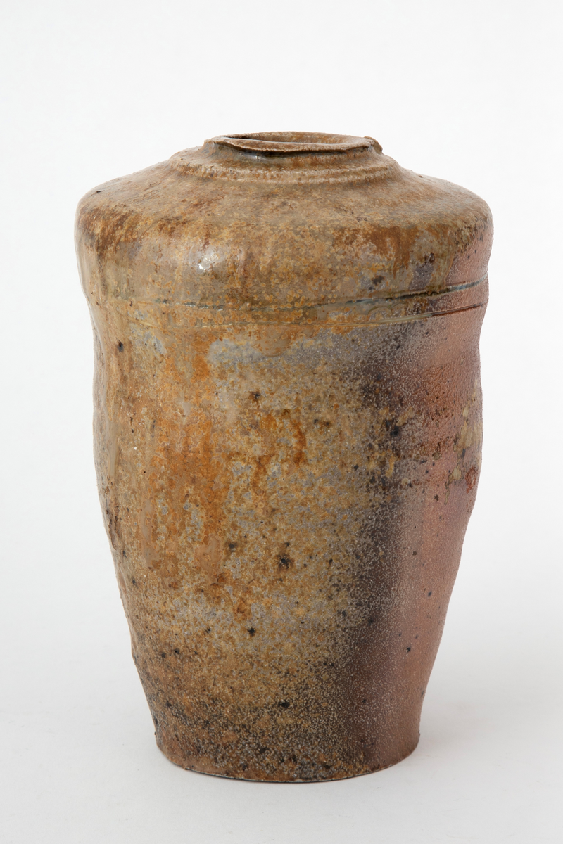 Krukke i steingods, avsmalnende nedover med uregelmessig profil. Liten åpning på svakt hvelvet, profilert topp. To ringer risset inn i godset på øvre del av korpus. Krukken er saltglasert og er brun på den ene siden og gulbrun på den andre.