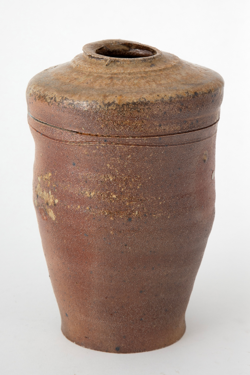 Krukke i steingods, avsmalnende nedover med uregelmessig profil. Liten åpning på svakt hvelvet, profilert topp. To ringer risset inn i godset på øvre del av korpus. Krukken er saltglasert og er brun på den ene siden og gulbrun på den andre.